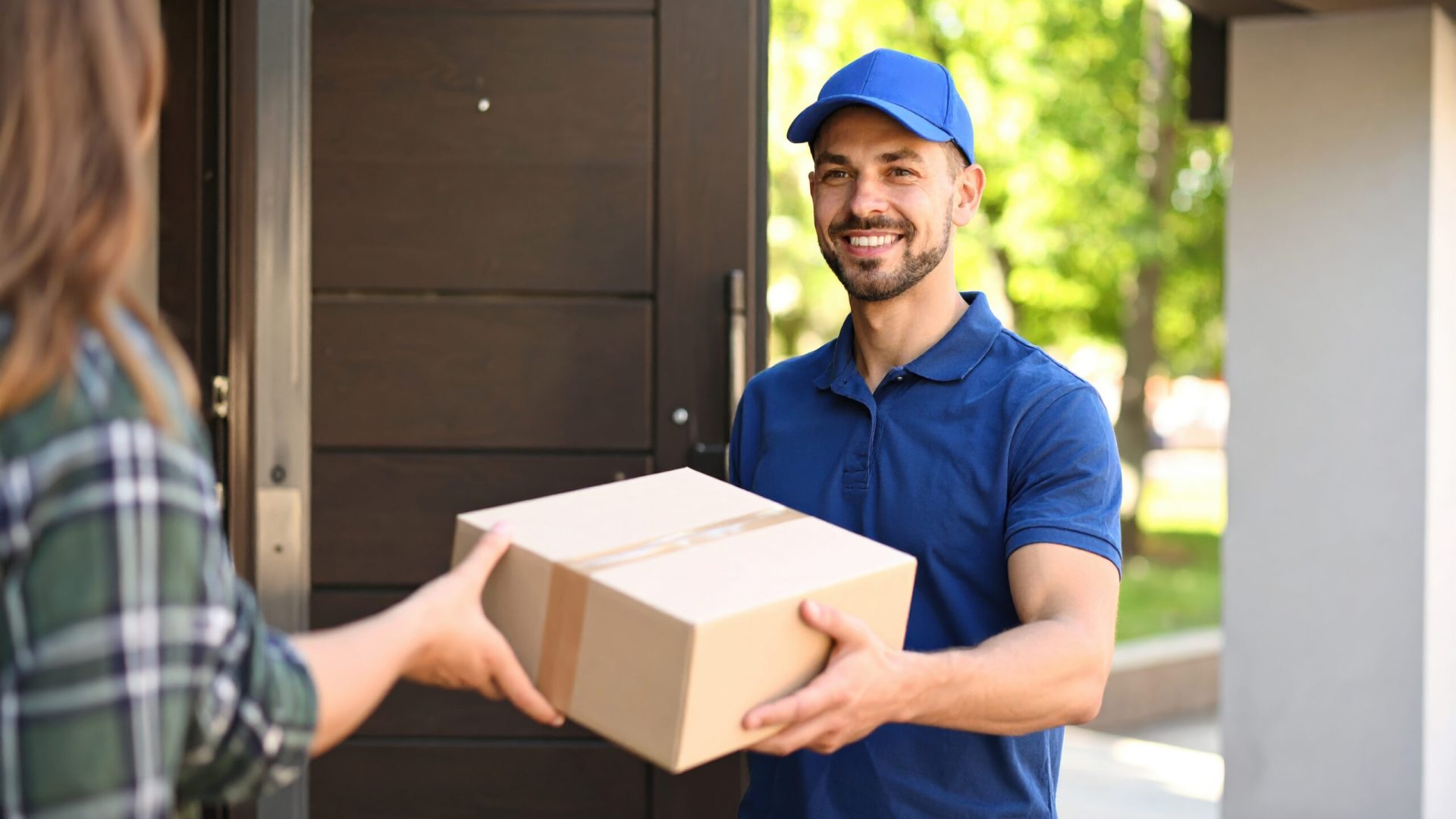 Delivery person in blue uniform handing over package to customer at doorstep on sunny day, smiling and showing friendly service, with background of green trees and modern house