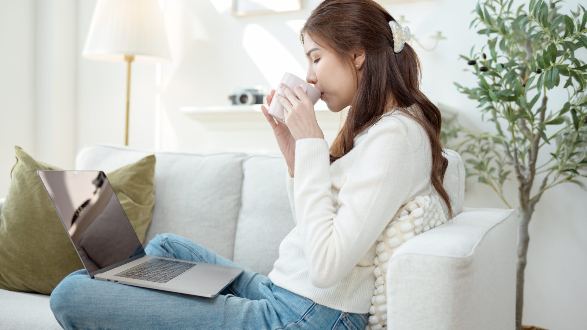 A young woman is focused on her laptop, holding a cup of coffee in a light-filled living room.